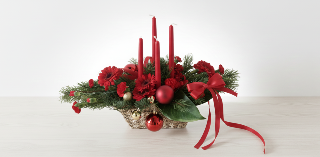 En modern juldekoration i en flätad korg med fyra röda ljus, röda blommor, granris och julkulor på en ljus träyta mot en vit bakgrund.