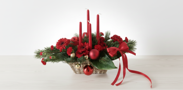 En modern juldekoration i en flätad korg med fyra röda ljus, röda blommor, granris och julkulor på en ljus träyta mot en vit bakgrund.